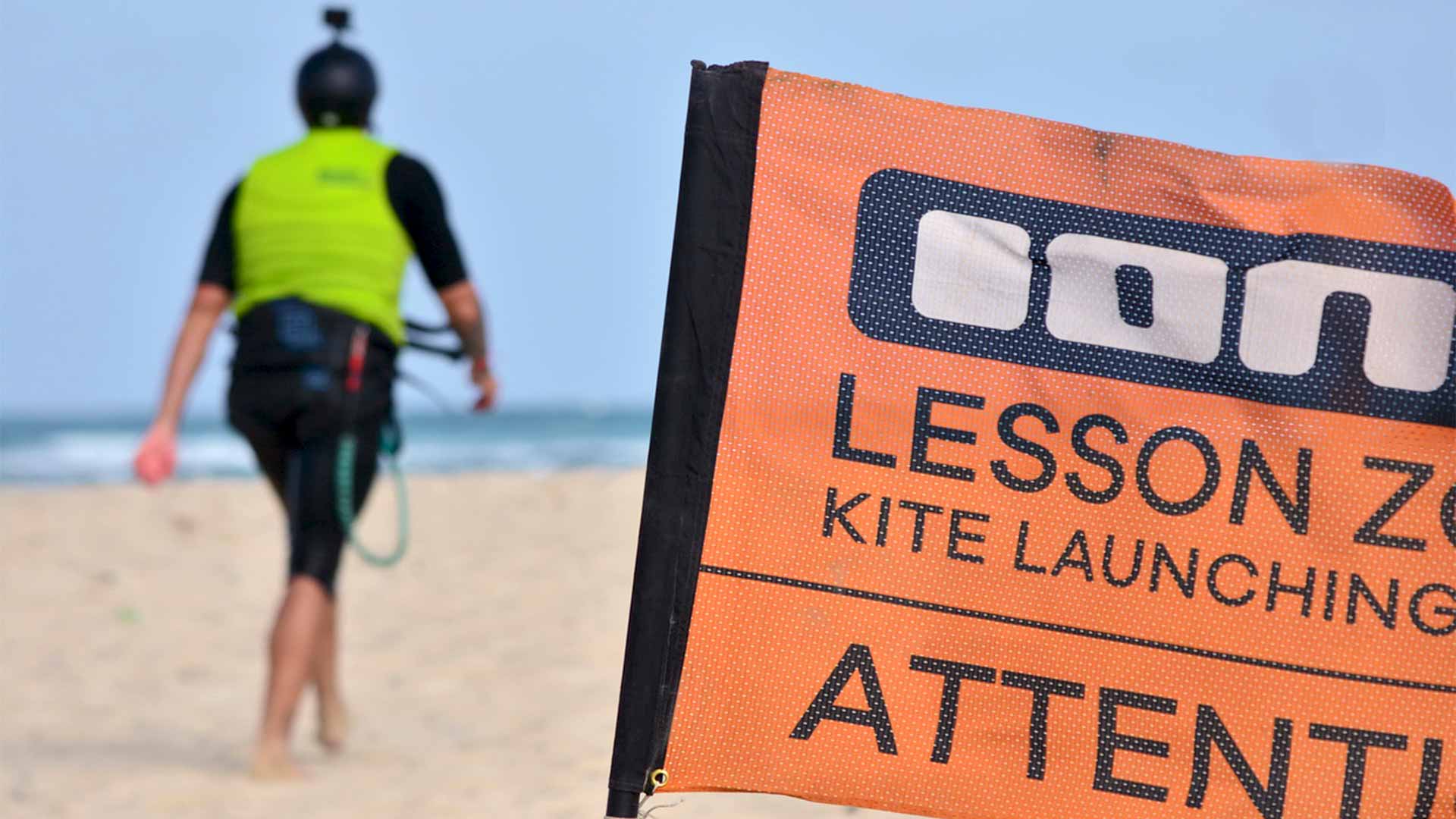 Devant, un drapeau indiquant la zone de cours de kitesurf et au loin, un étudiant marchant en tenue de sécurité.