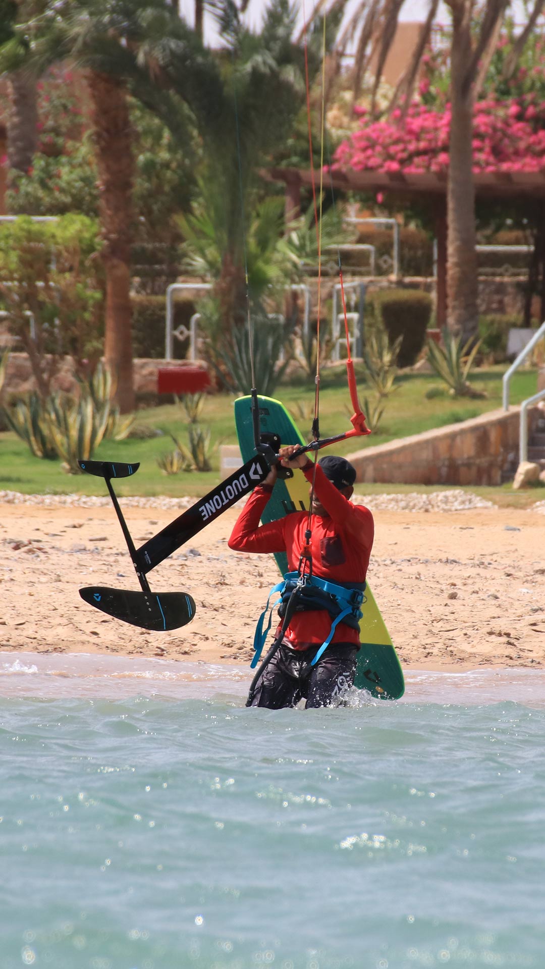Un instructor de kitefoil se prepara para navegar llevando la tabla de kitefoil con una mano y dirigiendo la cometa en el agua con la otra. 