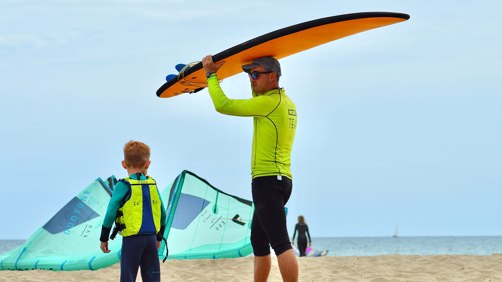 A surf instructor holds a beginner's surfboard above his head and is accompanied by a small boy for his first lesson in the water.