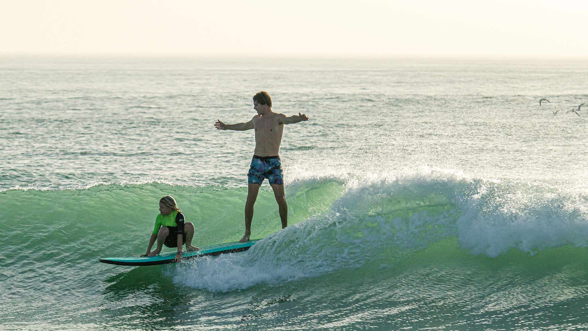 Father and son surfing a wave together on the same board, the boy crouching in front of the board and the father standing at the back.