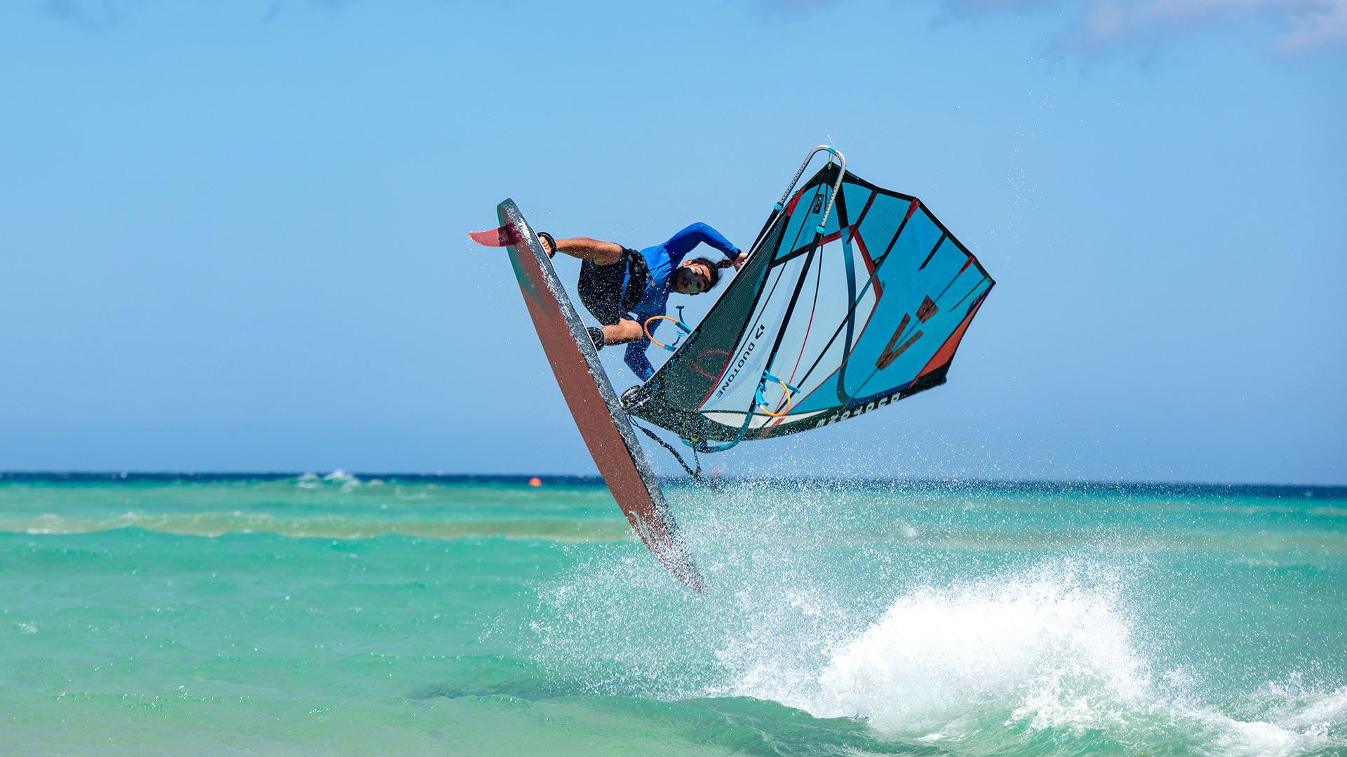 De windsurfinstructeur springt met het windsurfmateriaal op de Risco del Paso spot van Ion Club Fuerteventura, waar het heldere, turquoise water doet denken aan de sfeer van het eiland Maui. 