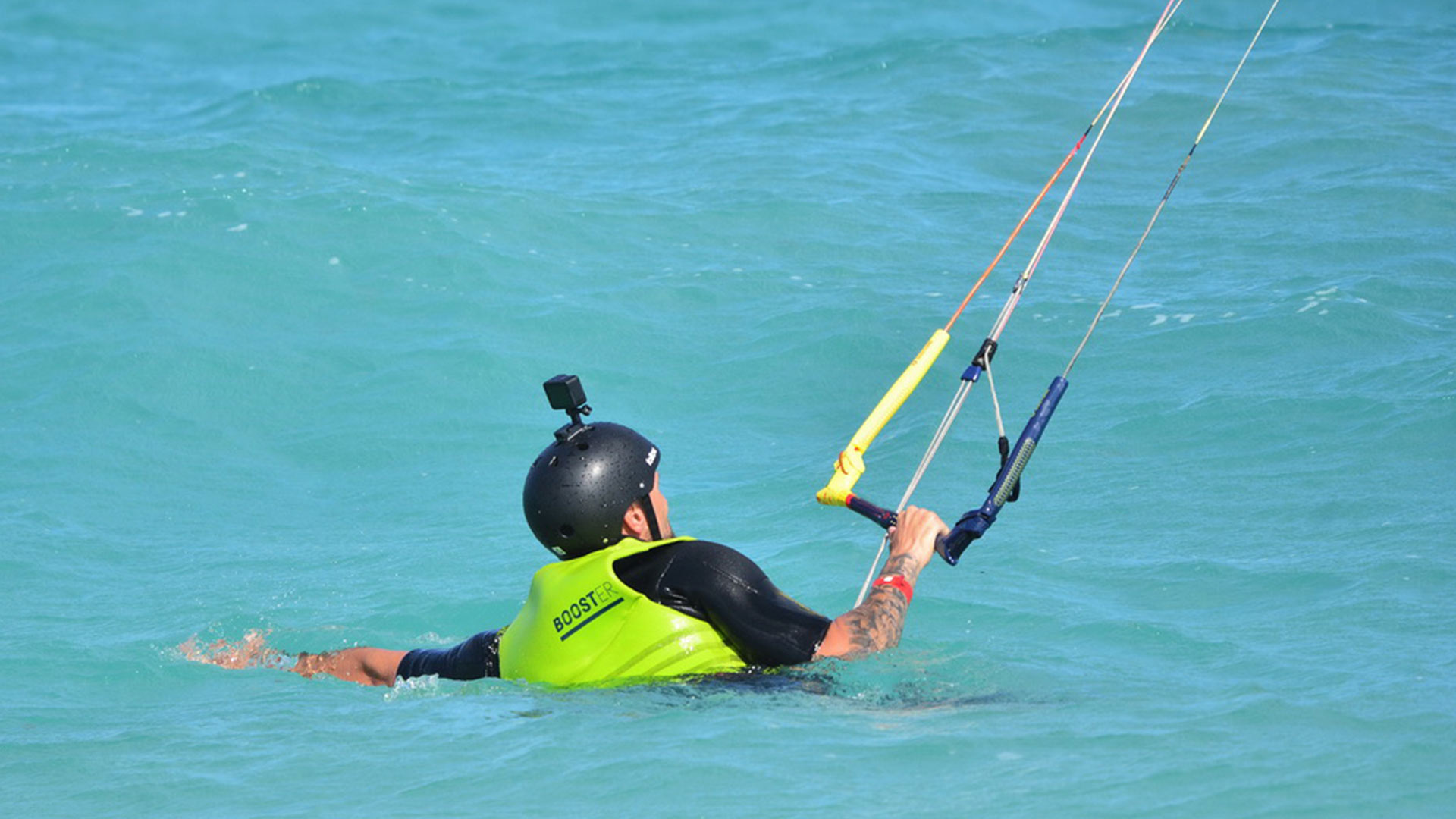 Secuencia de imágenes en las que un instructeur de kitesurf se cae realizando unos de los trucos para enseñárselo a sus estudiantes durante una clase   Devant un drapeau indiquant la zone de cours de kitesurf et au loin un étudiant marchant en tenue de sécurité.  Un instructeur enseignant comment attacher le leash au harnais de kitesurf, un étudiant du cours de kitesurf tenant un cerf-volant et un instructeur tenant une planche de kitesurf afin d'entrer dans l'eau pour naviguer.  Un étudiant du cours de kitesurf est au niveau 2, nageant dans l'eau en même temps qu'il contrôle son cerf-volant. 