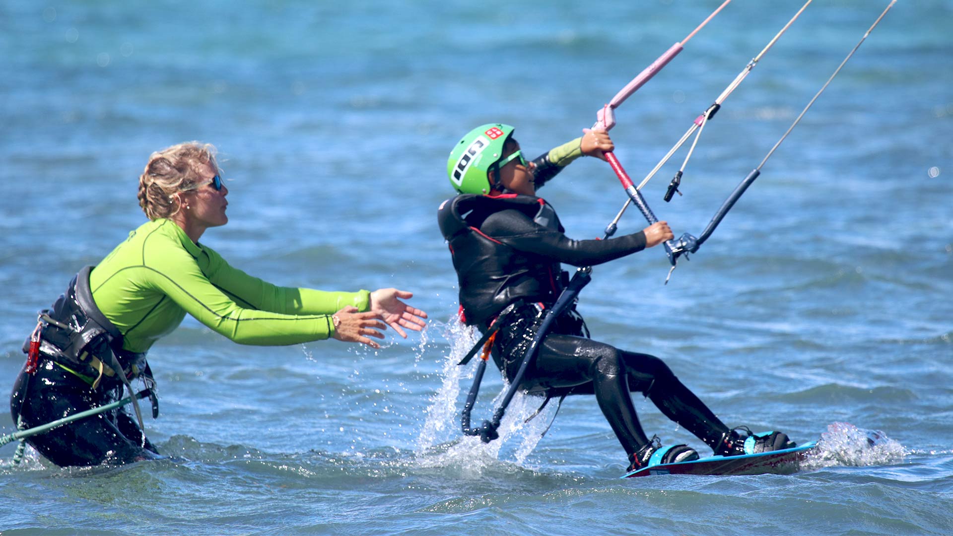 Secuencia de imágenes en las que un instructeur de kitesurf se cae realizando unos de los trucos para enseñárselo a sus estudiantes durante una clase   Devant un drapeau indiquant la zone de cours de kitesurf et au loin un étudiant marchant en tenue de sécurité.  Un instructeur enseignant comment attacher le leash au harnais de kitesurf, un étudiant du cours de kitesurf tenant un cerf-volant et un instructeur tenant une planche de kitesurf afin d'entrer dans l'eau pour naviguer.  Un étudiant du cours de kitesurf est au niveau 2, nageant dans l'eau en même temps qu'il contrôle son cerf-volant.   Un instructeur de kitesurf de Ion Club Centers enseigne le water star à sa petite fille. 