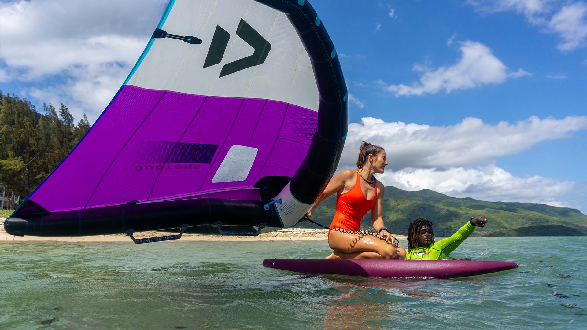 Un instructor de kitefoil se prepara para navegar llevando la tabla de kitefoil con una mano y dirigiendo la cometa en el agua con la otra. 